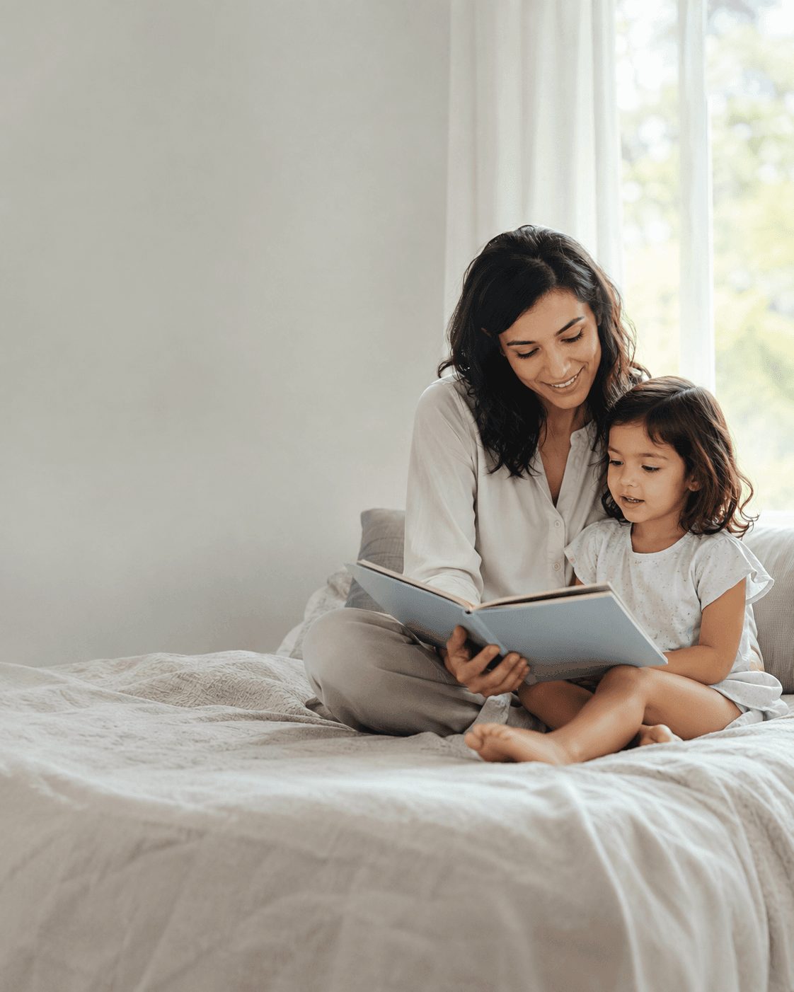 Mãe e filha lendo um livro juntas, sentadas na cama, em um quarto com luz natural.